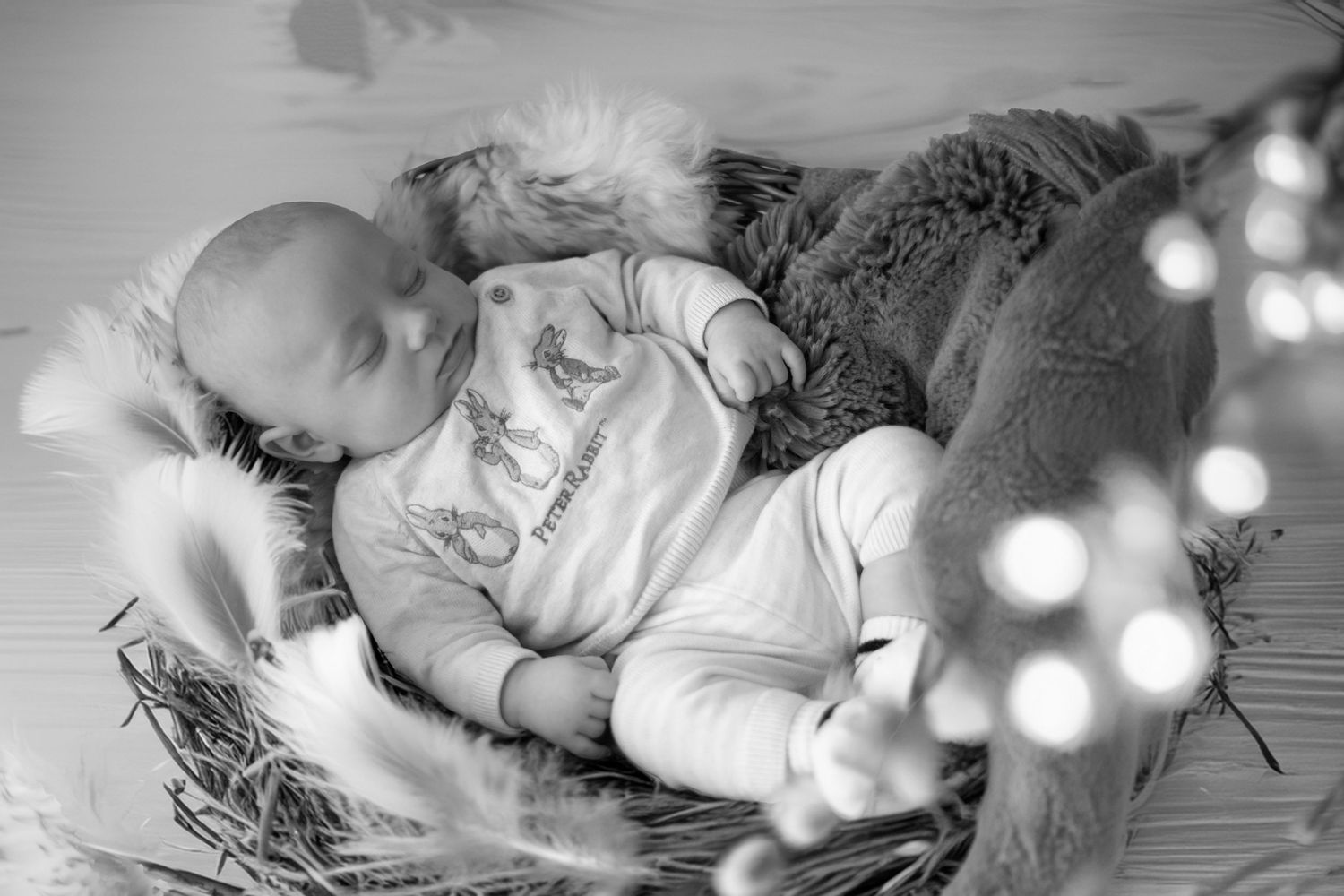Black and white photograph of a sleeping baby in white clothing nestled in a cozy basket with bokeh lights in background.