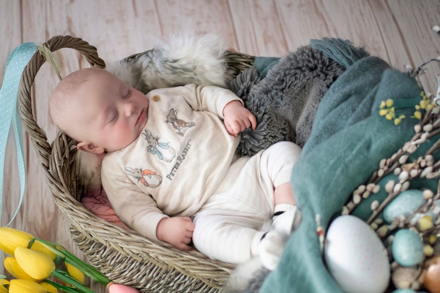 Baby sleeping peacefully in wicker basket with teal blanket and spring decor including yellow tulips and pussy willows.