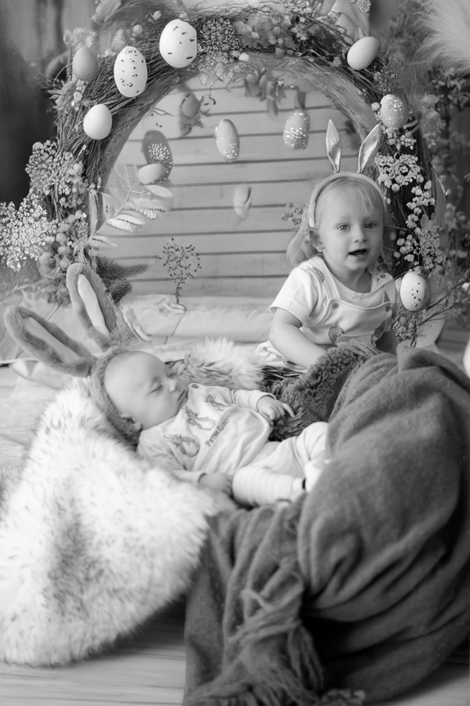 Black and white Easter photo with decorated arch, eggs, and children sitting among plush bunnies and decorative props.