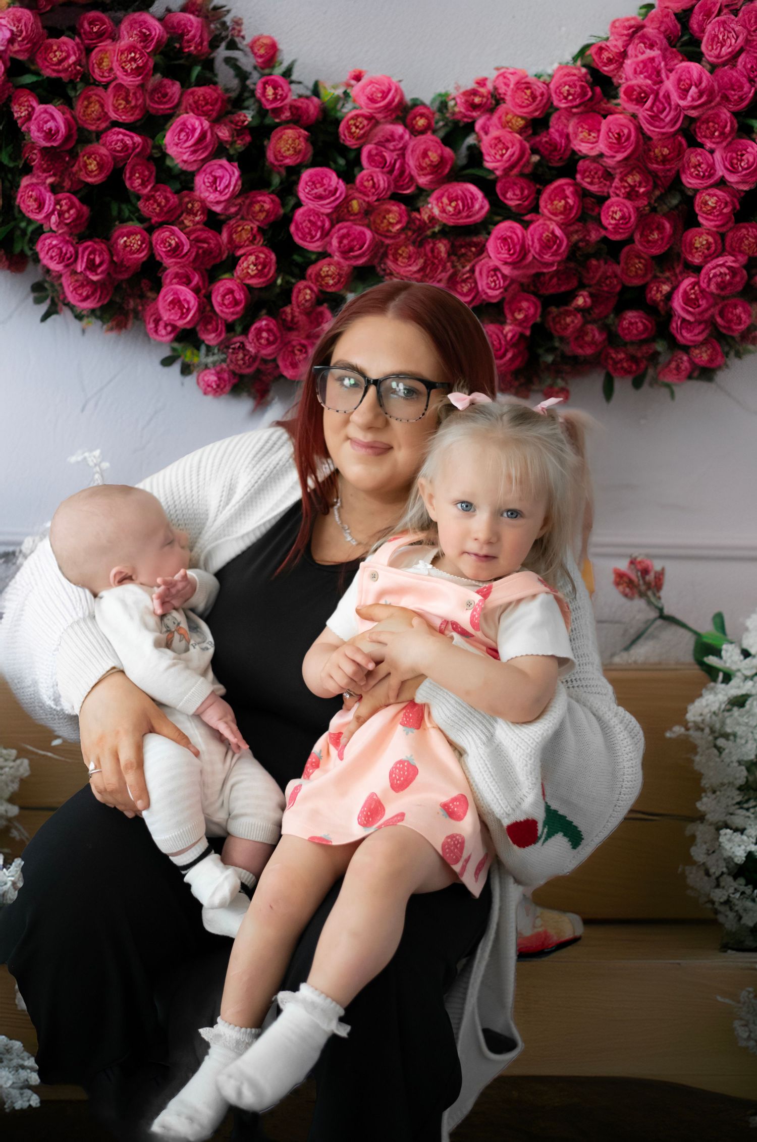 A family portrait against a wall of vibrant pink roses, featuring a small baby, toddler in pink, and an adult in glasses.