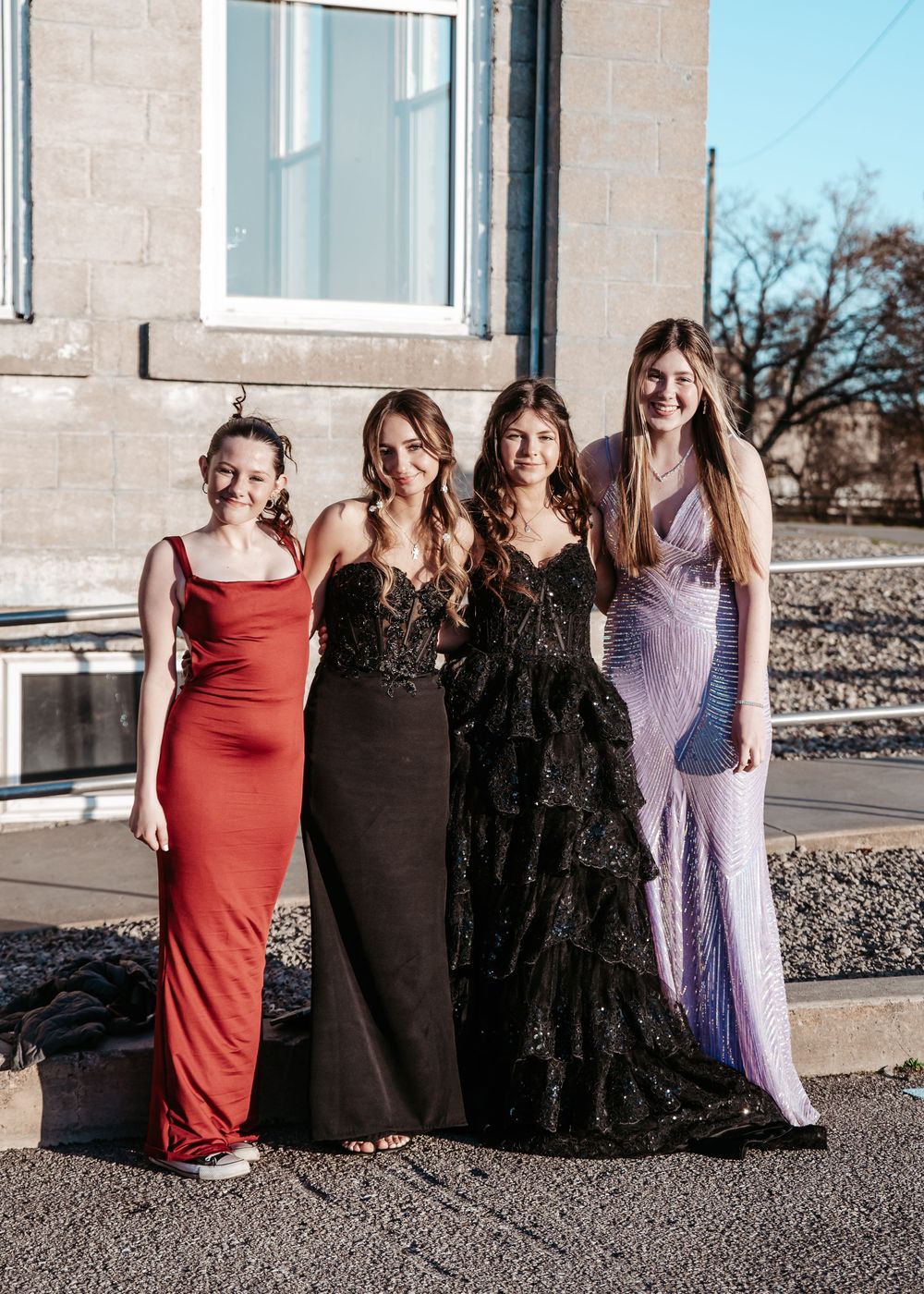 Four women stand together in elegant formal gowns including red, black and lavender dresses at an outdoor event.