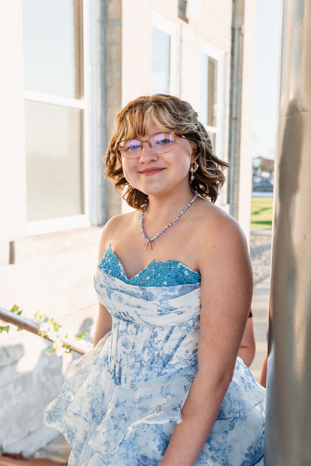 A person wearing a strapless blue and white patterned dress poses by white pillars in bright sunlight.