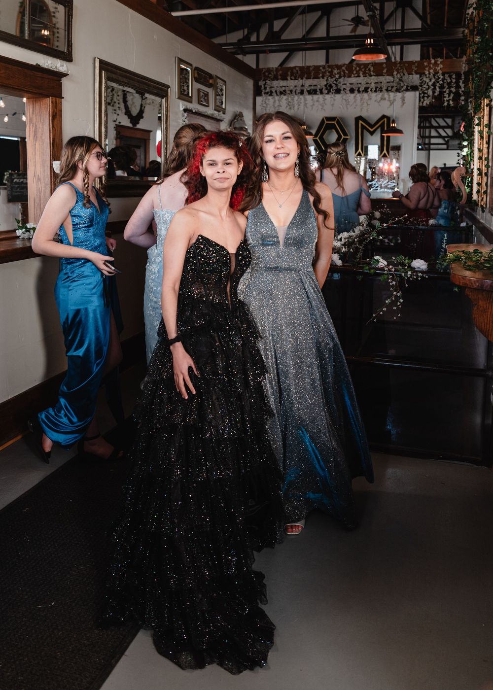 Two people in elegant formal dresses pose together at a party venue with a black sparkly gown and silver glittery dress.