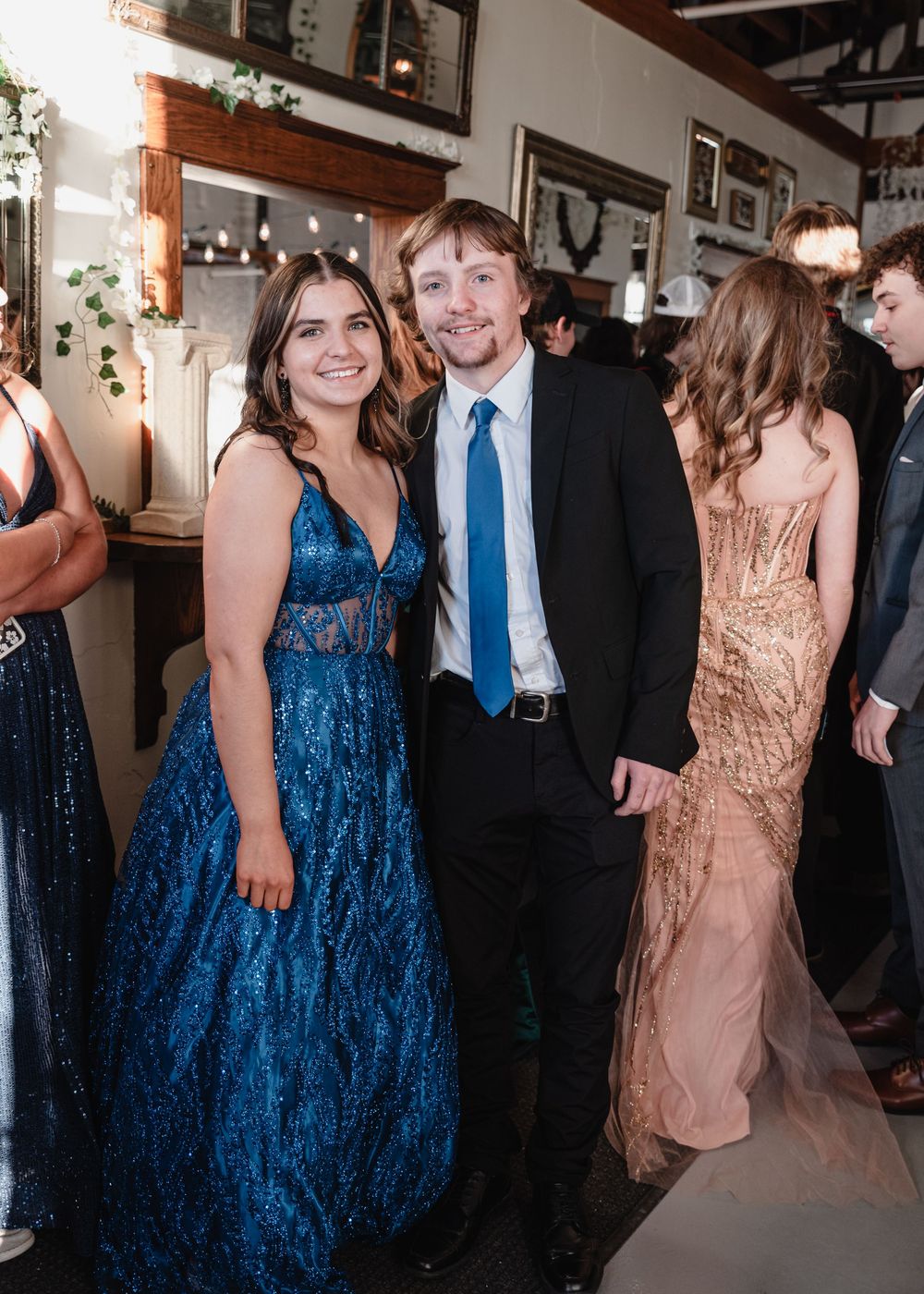 A couple dressed in formalwear attend an event with others in a rustic indoor venue decorated with photo frames.