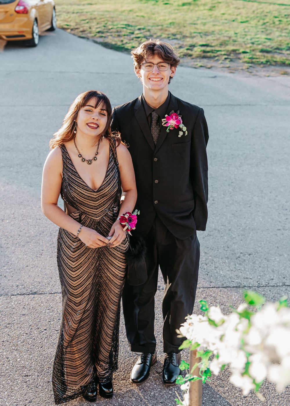 Two people in formal attire stand together on a paved road, with the person in a black suit wearing a pink floral boutonniere.