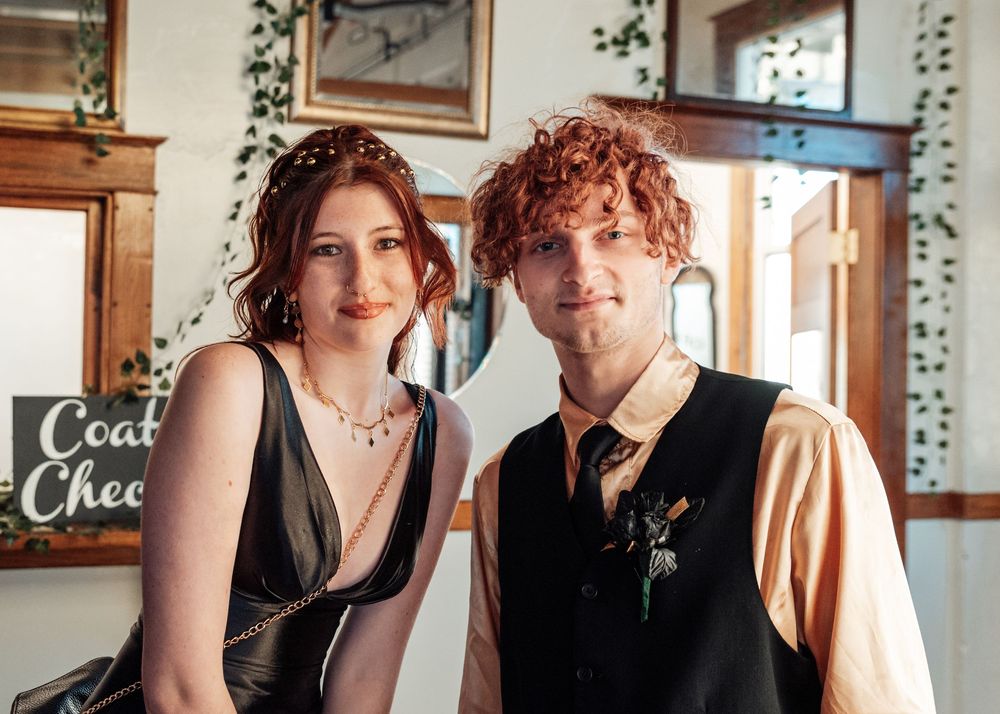 Two people dressed in formal vintage-inspired attire pose together at an indoor venue with greenery decor.