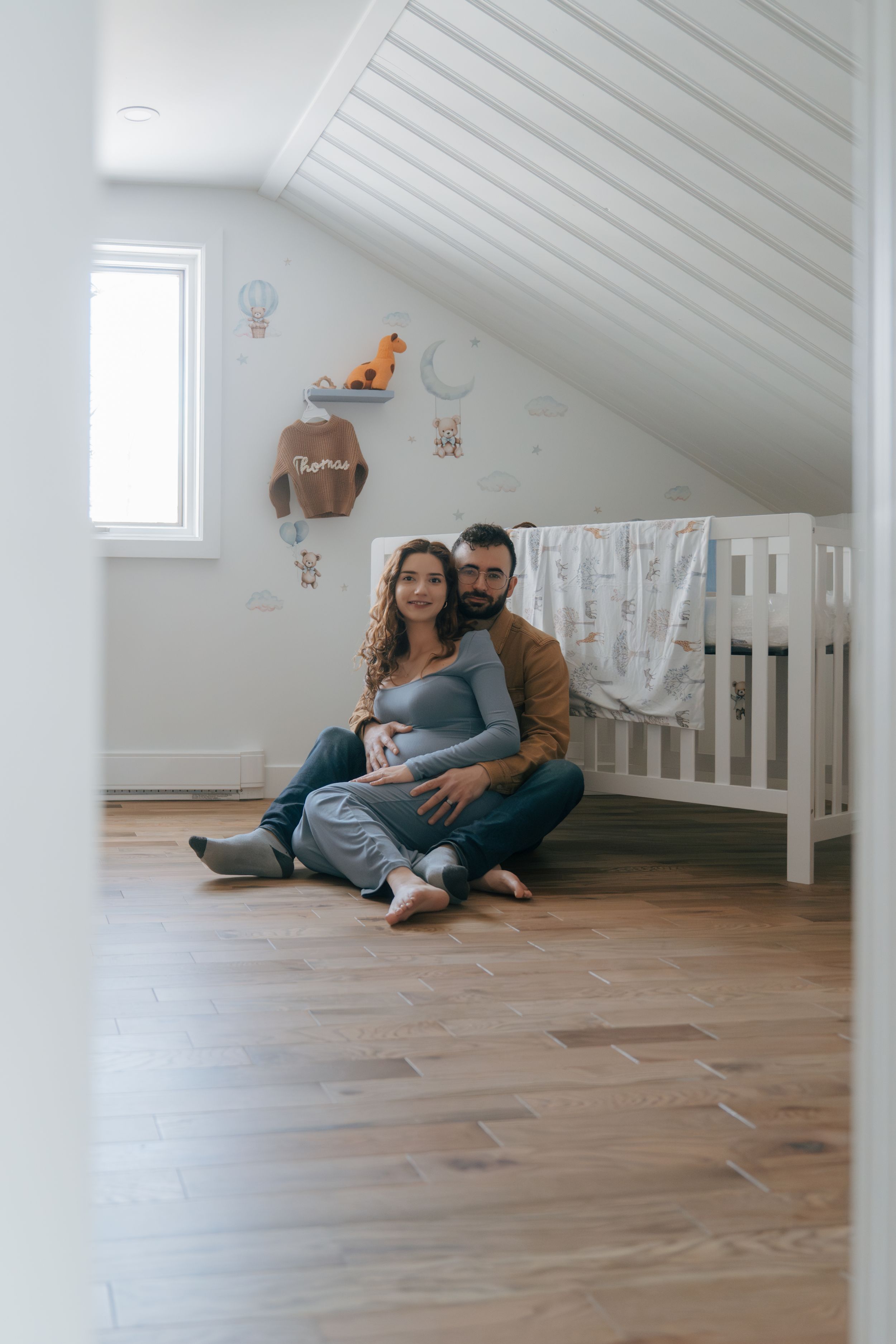 A cozy nursery room with a white crib, sloped ceiling, and couple sitting together on hardwood floor.