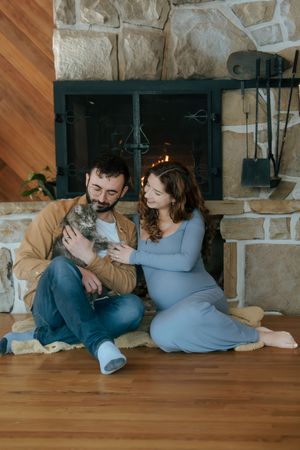 A couple relaxes by the fireplace with their cat, with the woman in a blue dress showing a pregnancy bump.