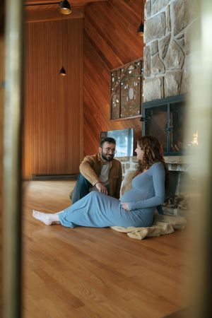 A couple relaxes together on a hardwood floor beside a stone fireplace in a cozy cabin interior.