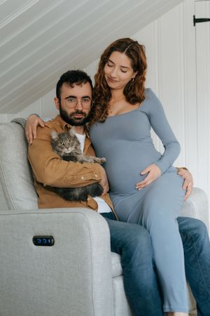 A couple relaxes on a gray couch with their fluffy gray cat as they await their new arrival together.