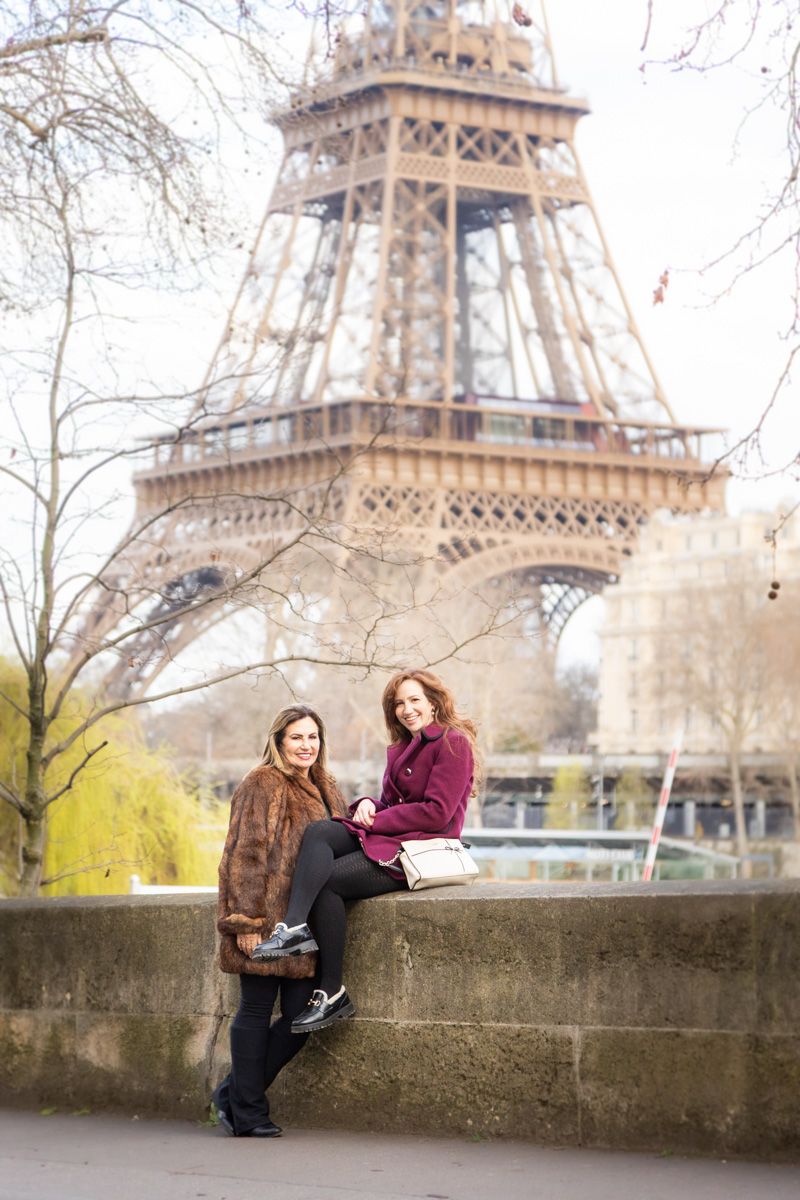 Two people sitting on stone wall near the Eiffel Tower in Paris on a winter day with bare trees in background.