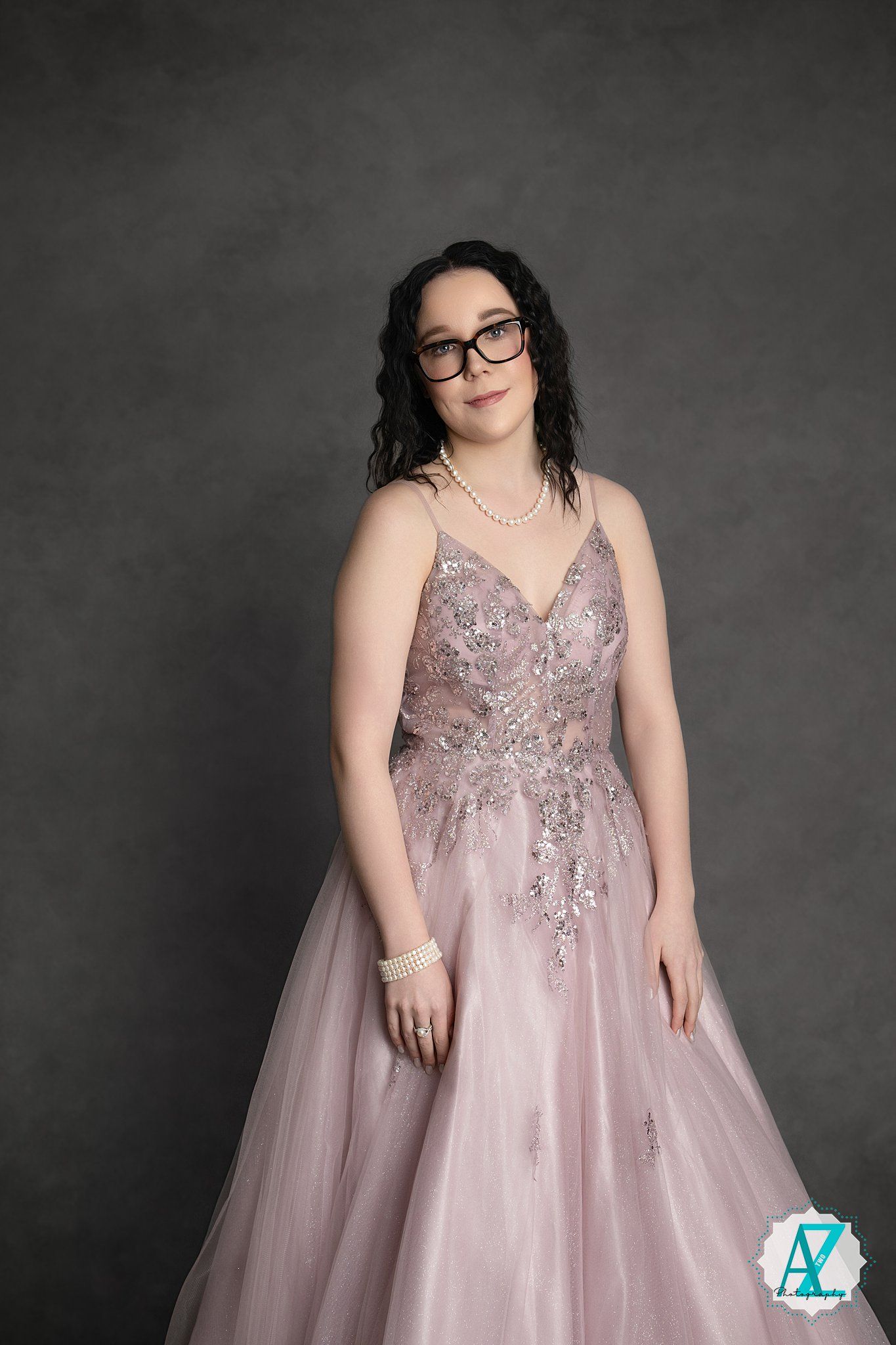 Person in glasses wearing an elegant pink tulle ball gown with beaded bodice against a dark gray backdrop.