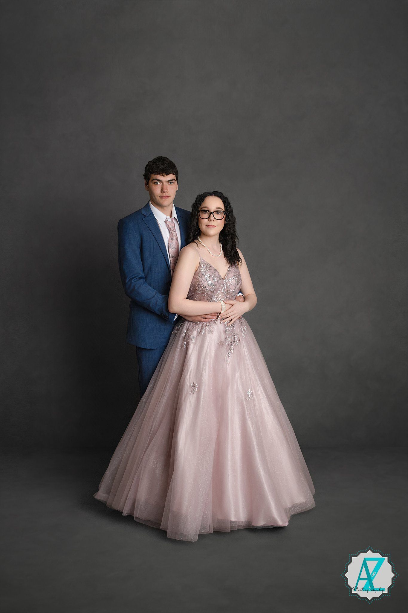 Formal couple poses in elegant attire with woman wearing pink tulle gown and man in blue suit against gray backdrop.