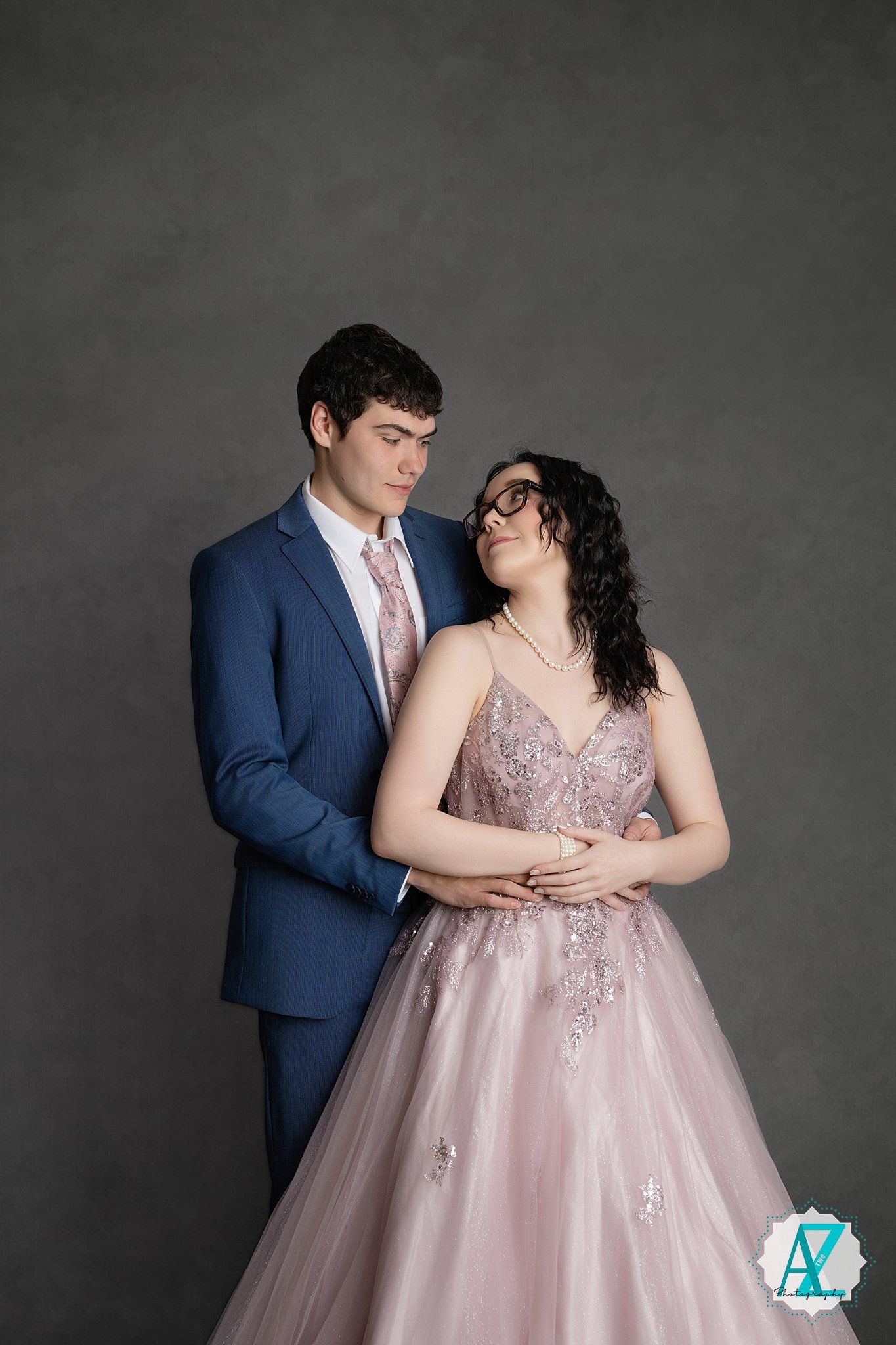 Romantic couple poses in formal attire, with navy suit and blush pink ballgown against dark gray backdrop.