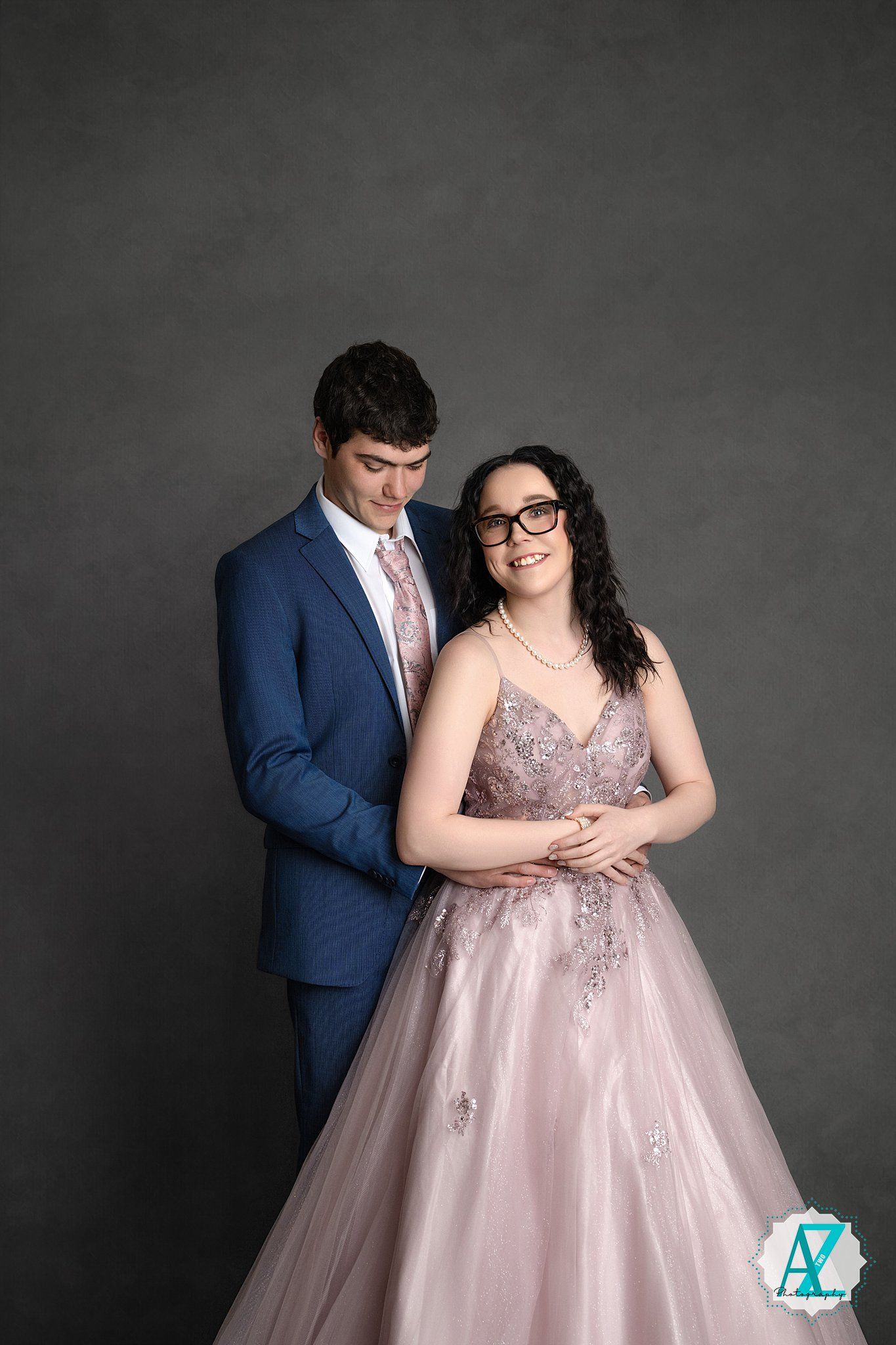 A formal couple poses against a gray backdrop, with her wearing a pink ballgown and him in a navy suit.