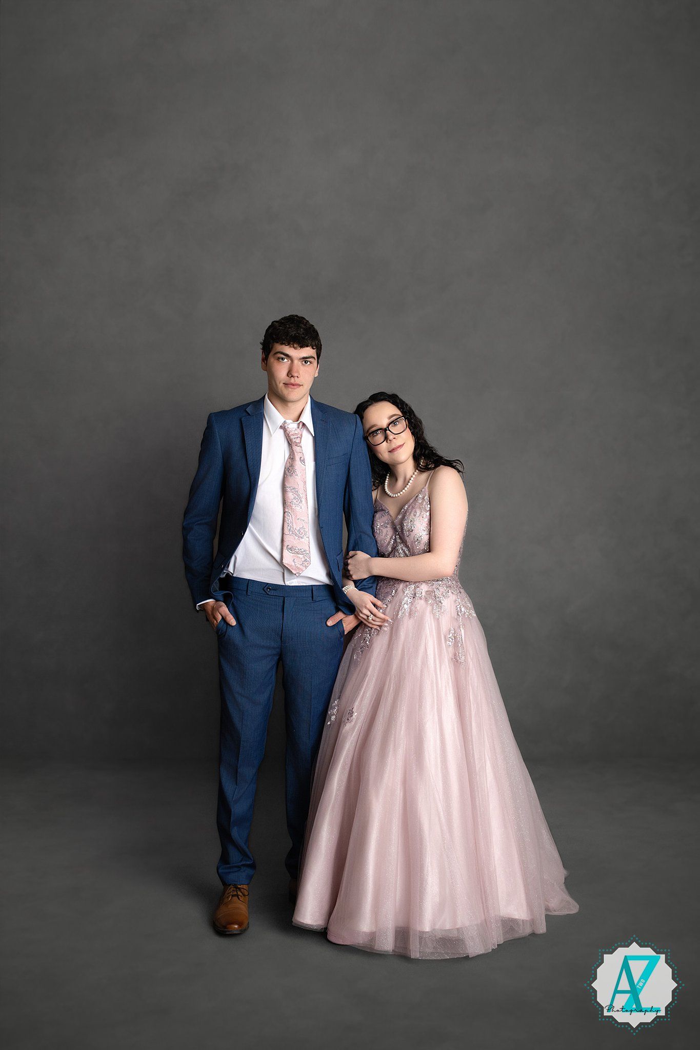 A couple in formal attire poses against a gray backdrop, with her in a pink tulle gown and him in a navy suit.