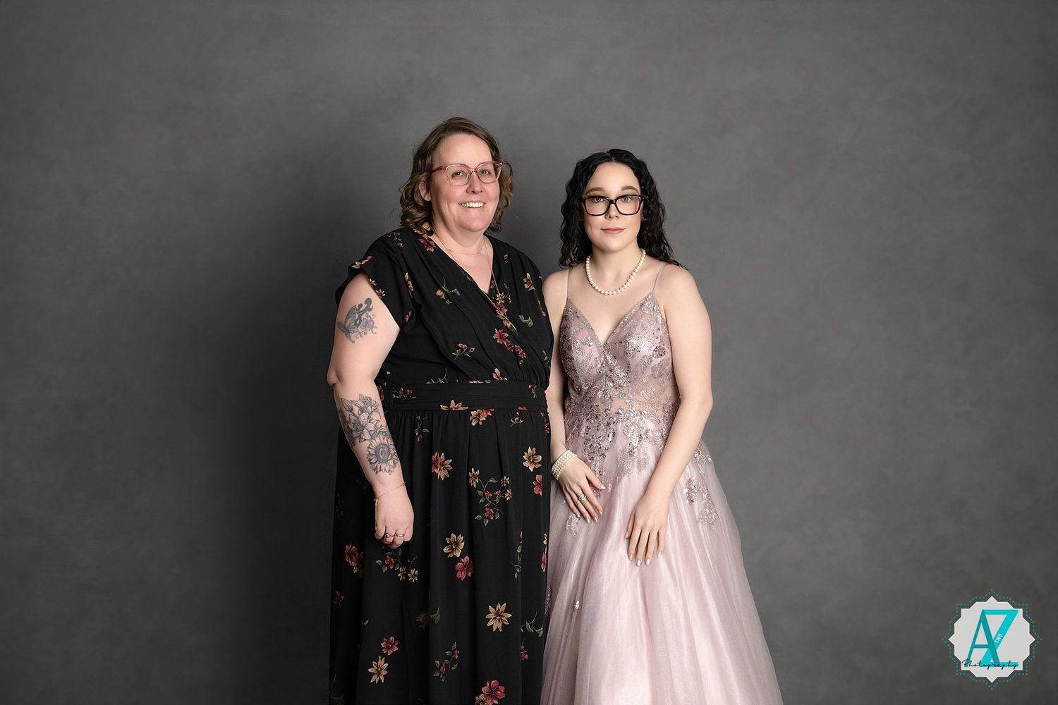 Two people pose for a formal portrait wearing a floral dress and a sparkly pink gown against a grey backdrop.