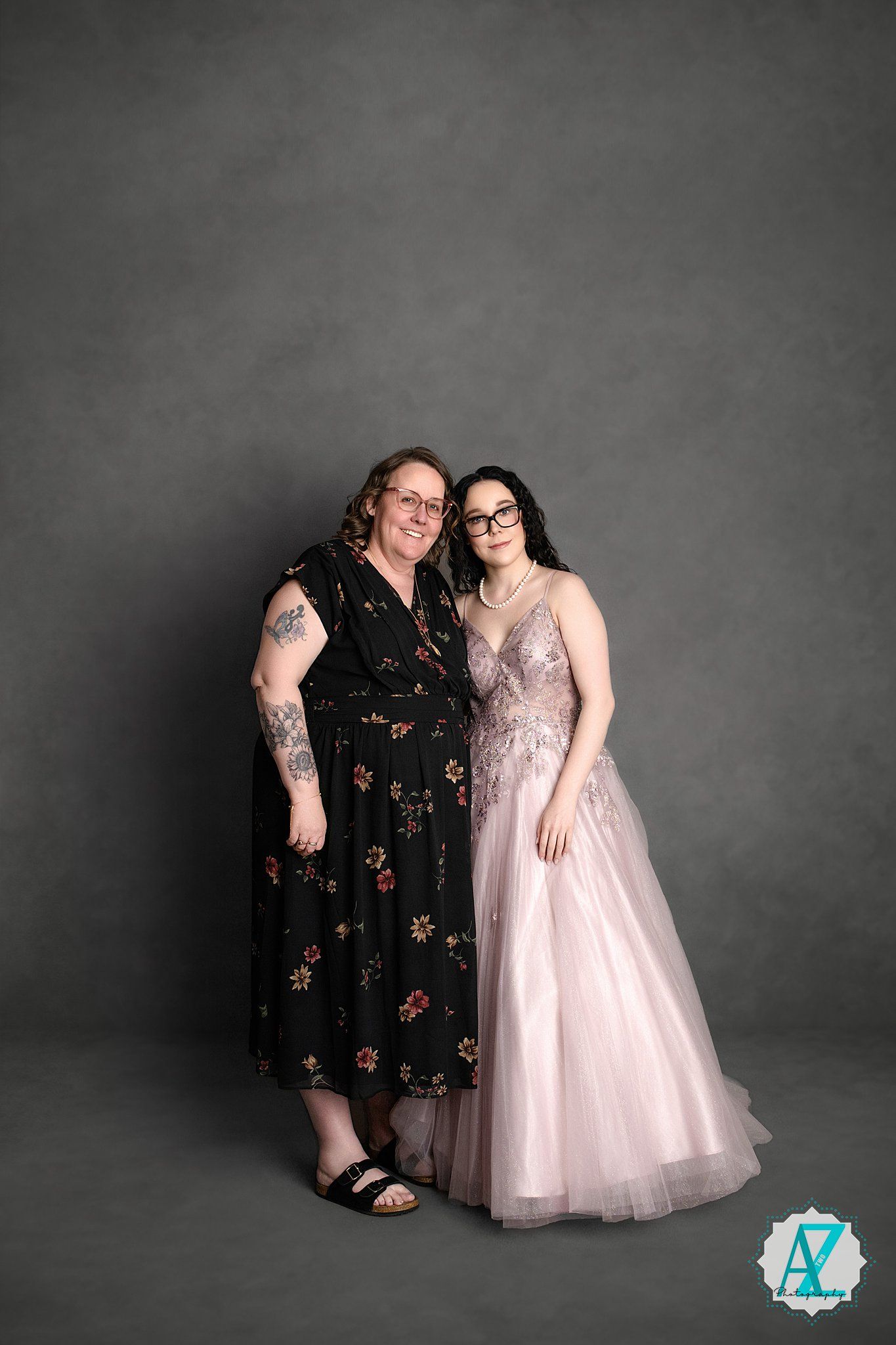 Two people pose together against a gray backdrop wearing a floral black dress and sparkly pink gown.