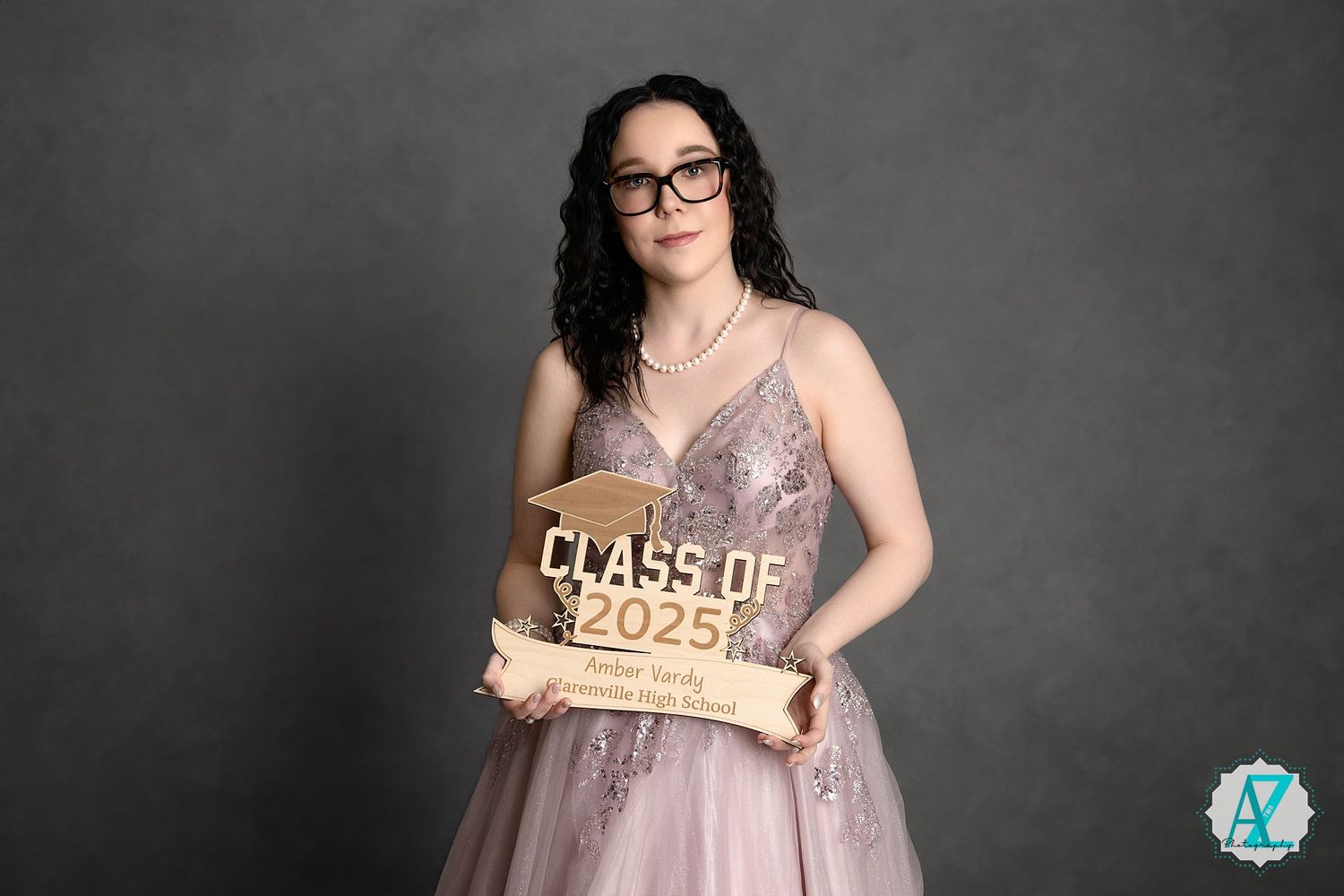Graduate in flowing mauve gown and glasses holds rustic wooden 'Class of 2023' sign against dark backdrop.