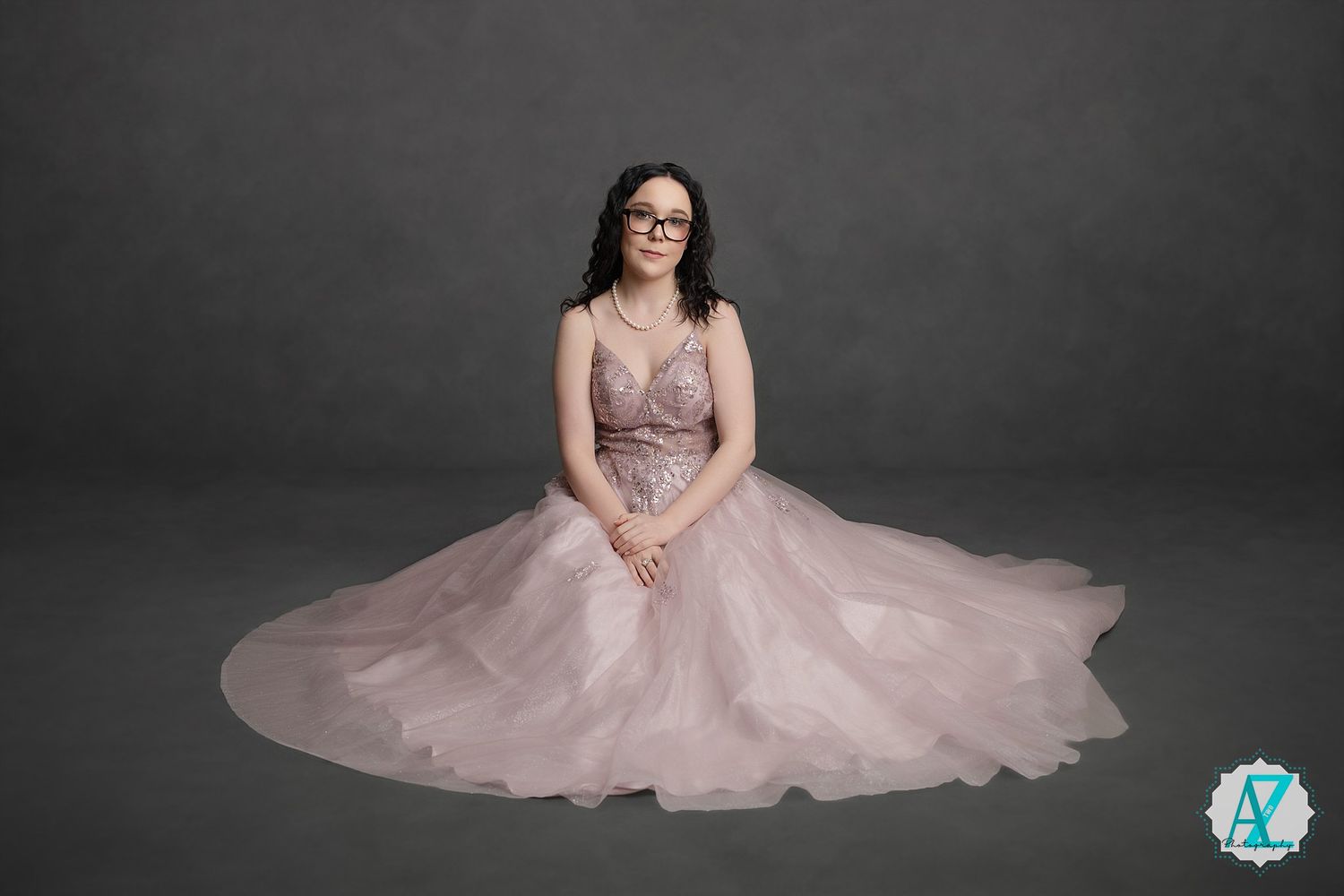A seated figure wearing a flowing pink tulle ballgown and glasses in a dramatic studio portrait against a dark background.