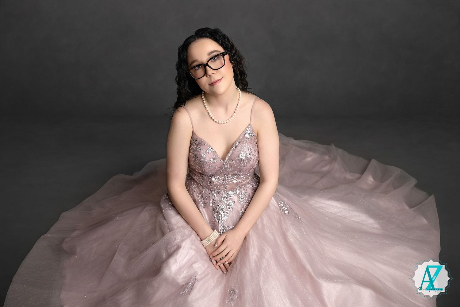 A model in glasses wearing a sparkly pink ballgown poses against a dark backdrop during a glamorous photoshoot.