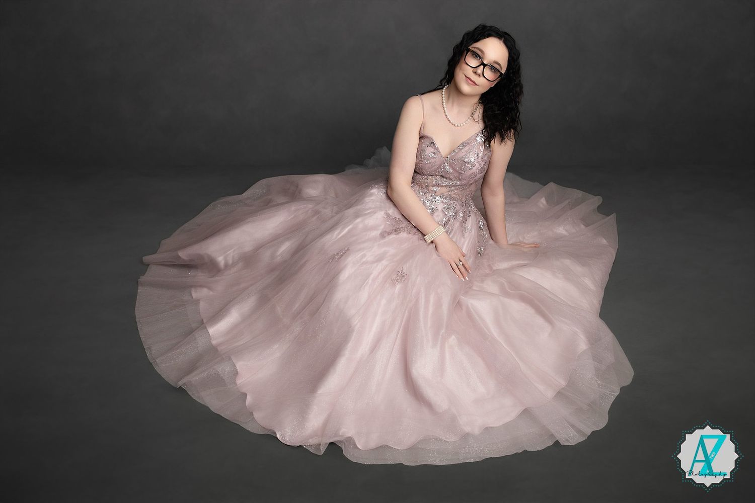 A figure sits elegantly in a flowing blush pink ball gown with sparkle details against a dark grey background.