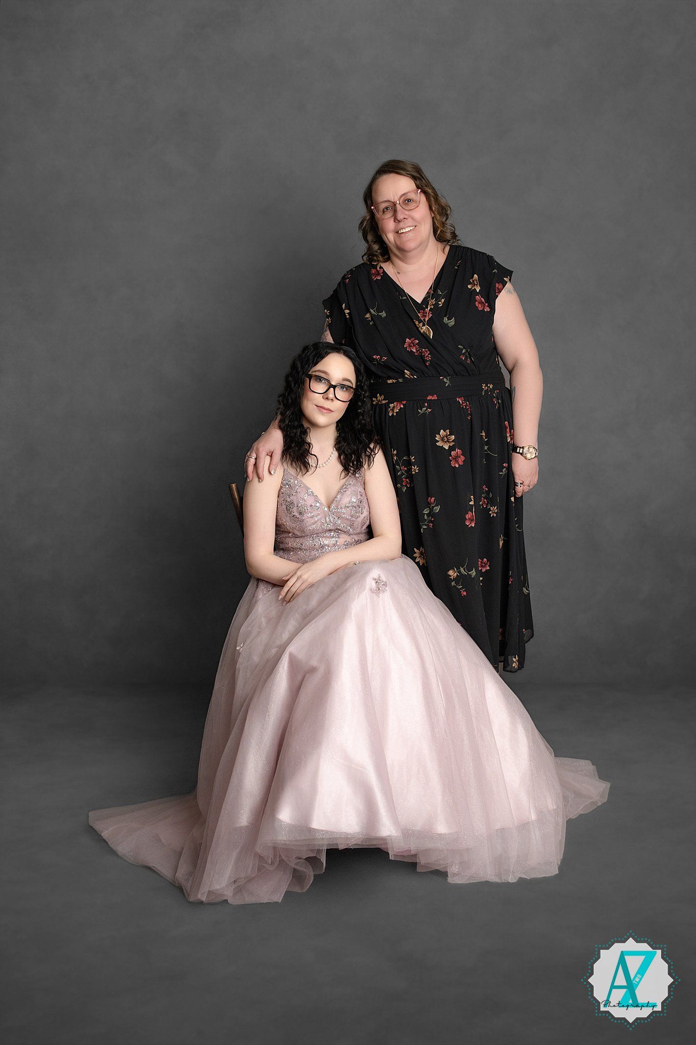 Studio portrait of two people posing, one seated in a pink ballgown and one standing in a floral dress against grey backdrop.