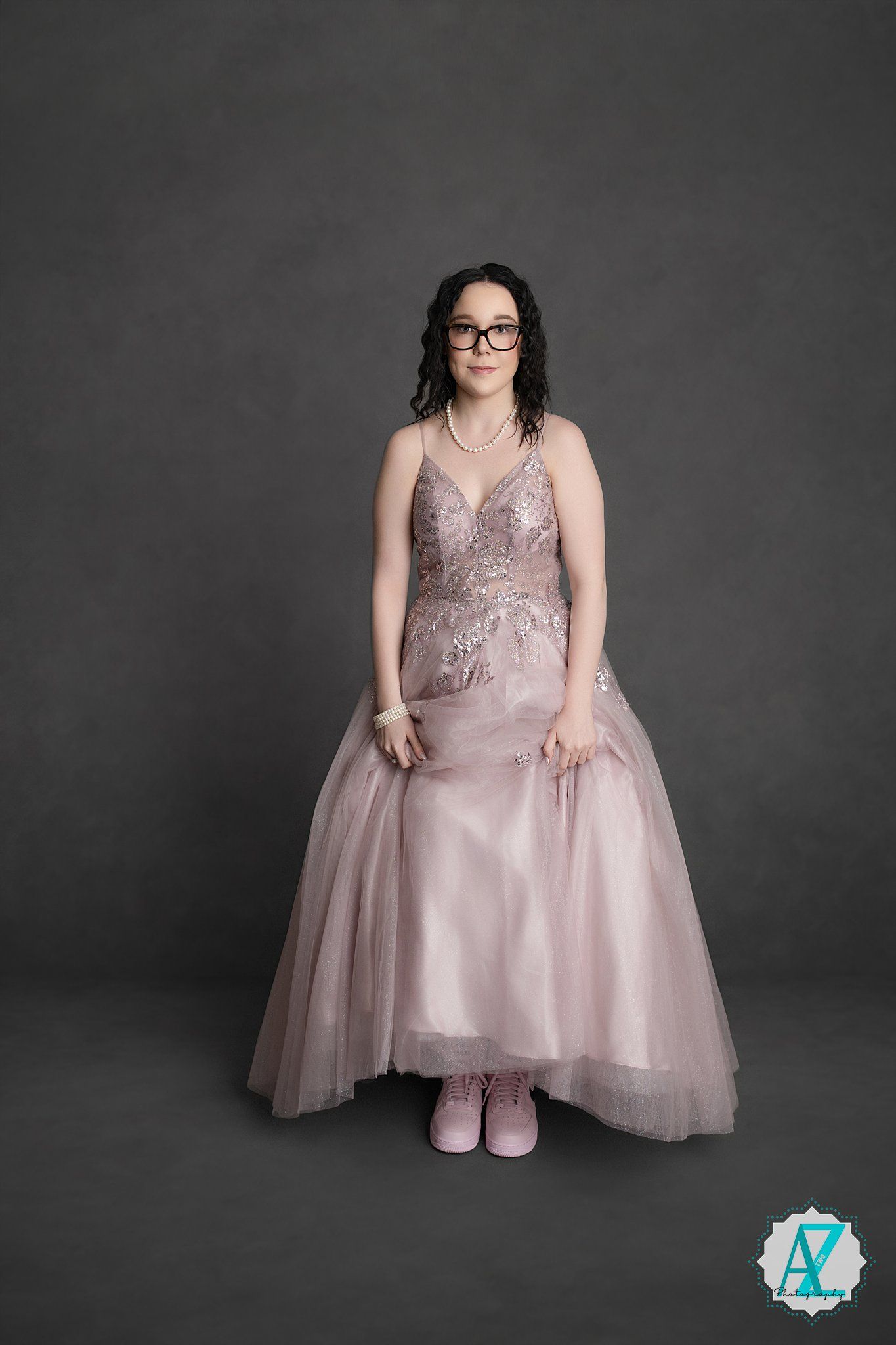 Person wearing glasses and a pale pink tulle ball gown with beaded bodice poses against a dark gray backdrop.