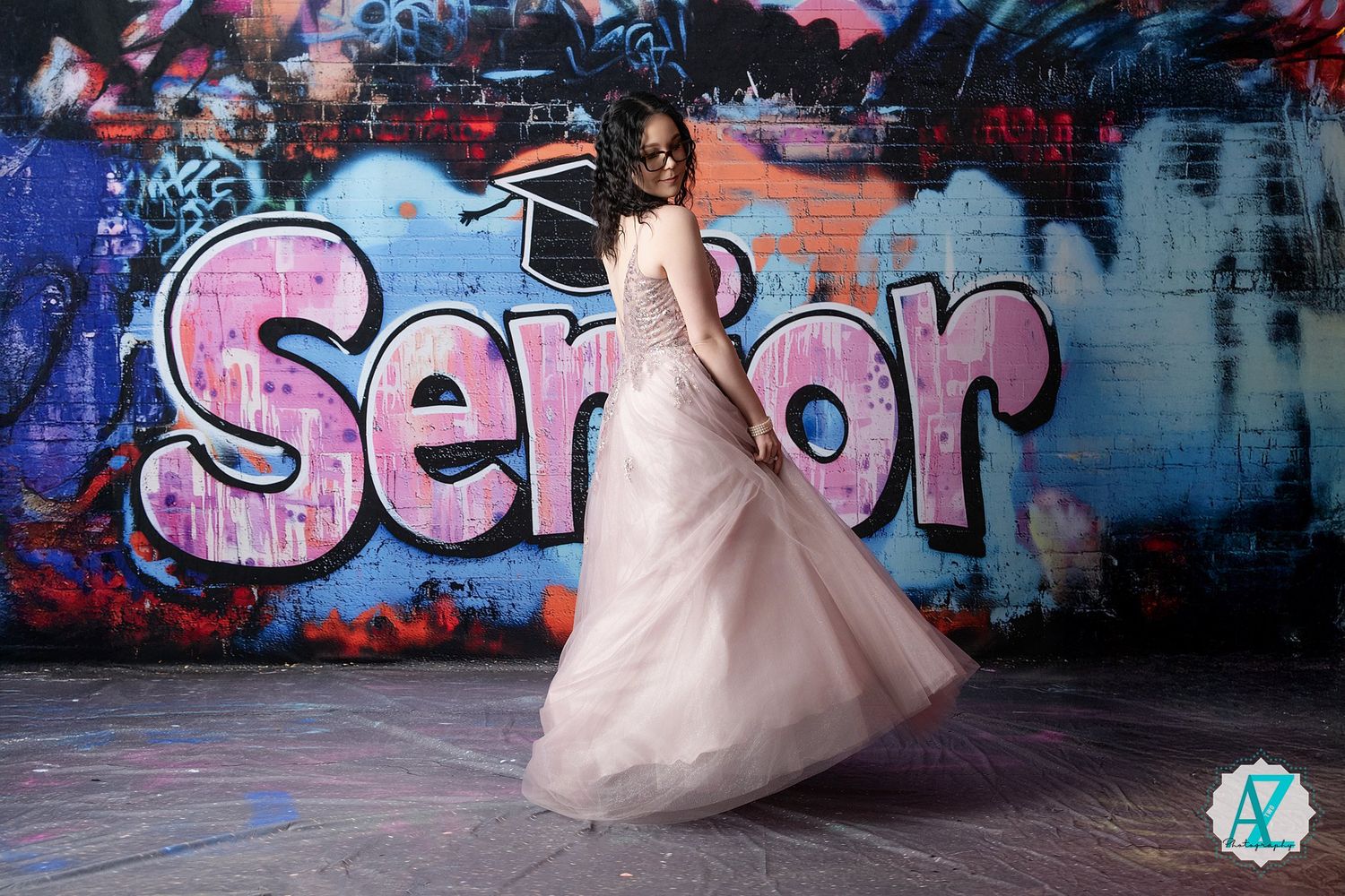 A person in a flowing pink tulle gown spins in front of vibrant graffiti art featuring the word 'Senior' in pink lettering.