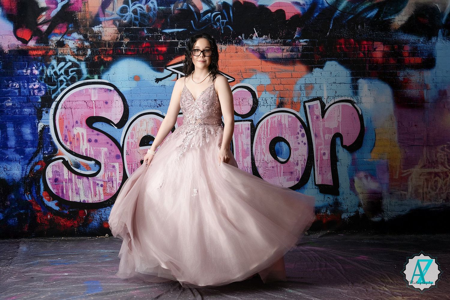 A person in a flowing blush pink formal gown twirls in front of a colorful graffiti wall with senior text.