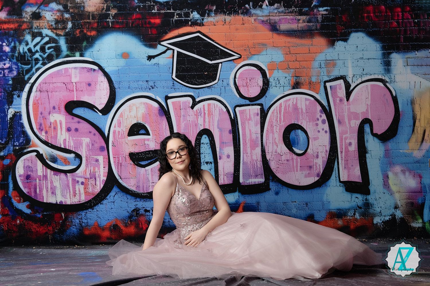 A formal senior photo featuring a pink tulle gown against a graffiti wall with "Senior" text and graduation cap artwork.
