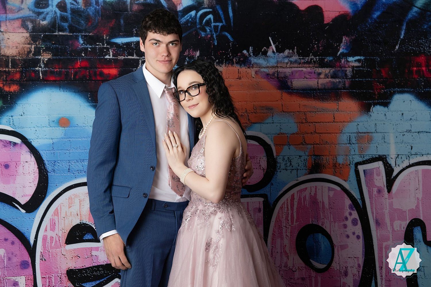 A couple poses together against a graffiti wall, wearing formal attire with a blue suit and pink dress.