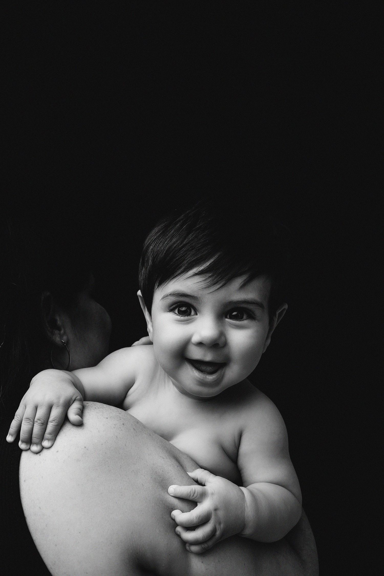Black and white portrait of a smiling baby being held against dark background showing pure joy and innocence.