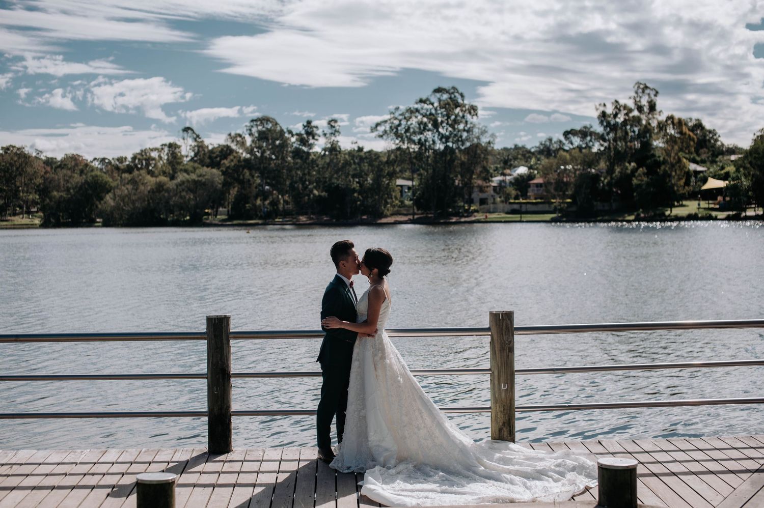 A couple in wedding attire embraces on a wooden dock overlooking a serene lake with trees and houses in the background.