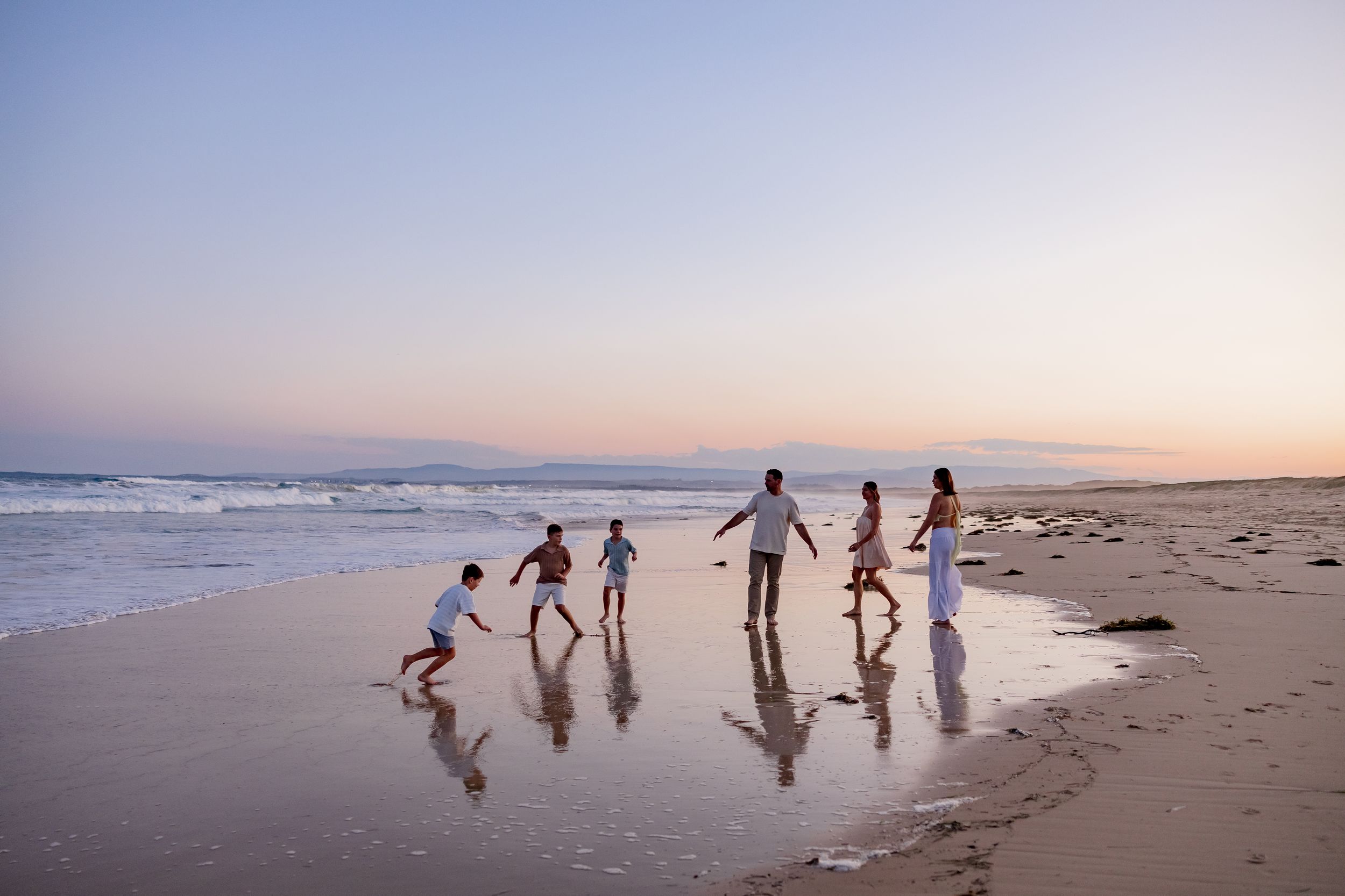 Silhouetted people running and playing together on a beach at sunset with waves and reflections in the wet sand.