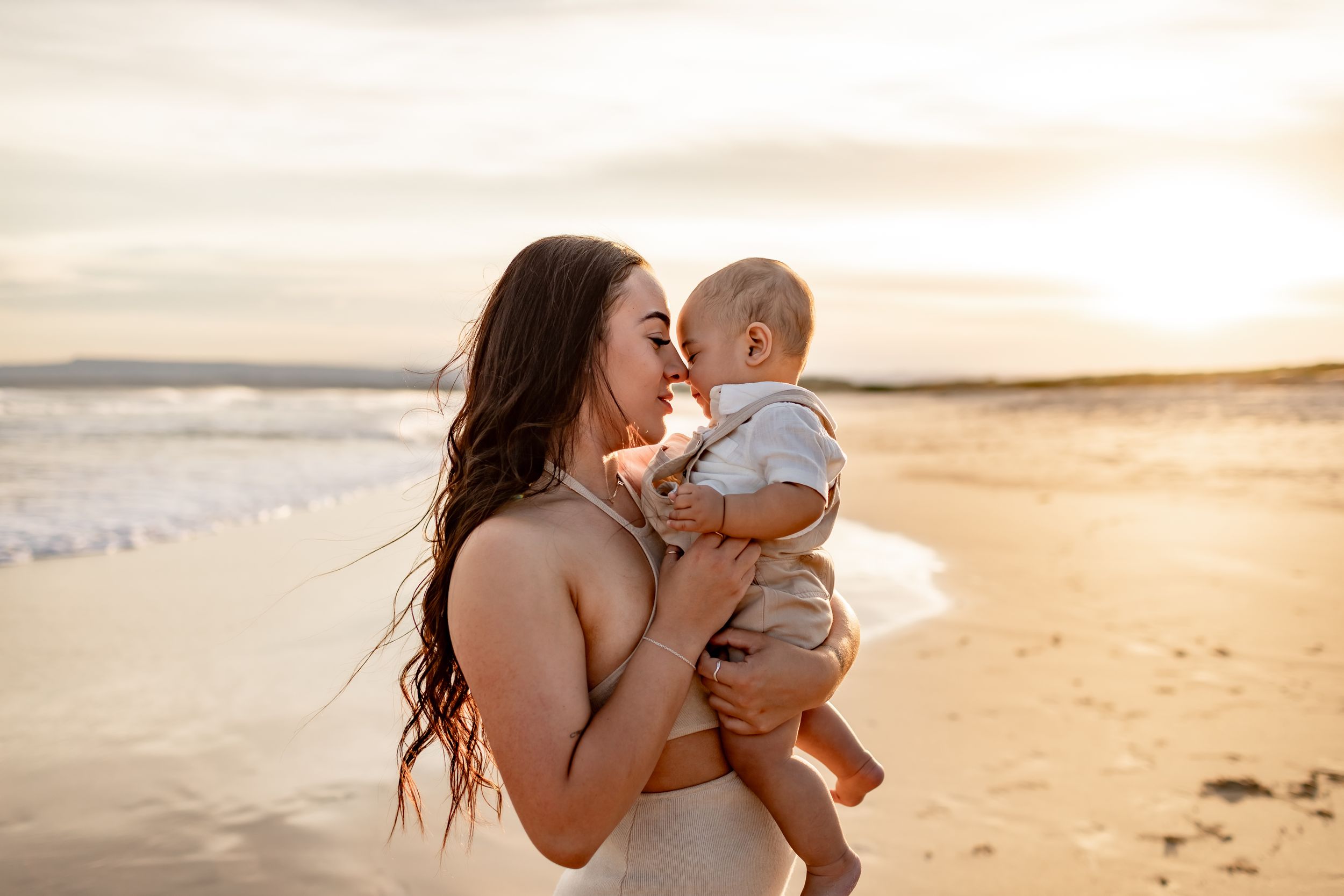 Tender moment between parent and baby against a glowing sunset beach backdrop.