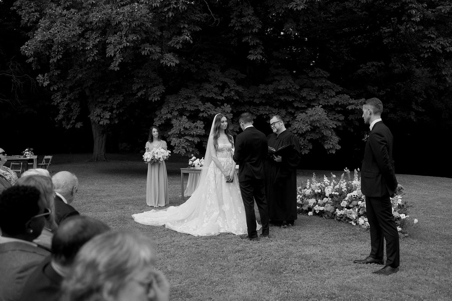 An outdoor wedding ceremony setup with guests gathered under trees.