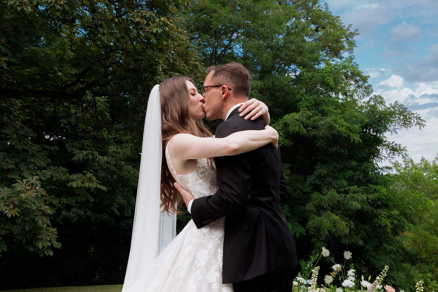 A romantic outdoor wedding moment of a first kiss captured among green trees and blue sky.