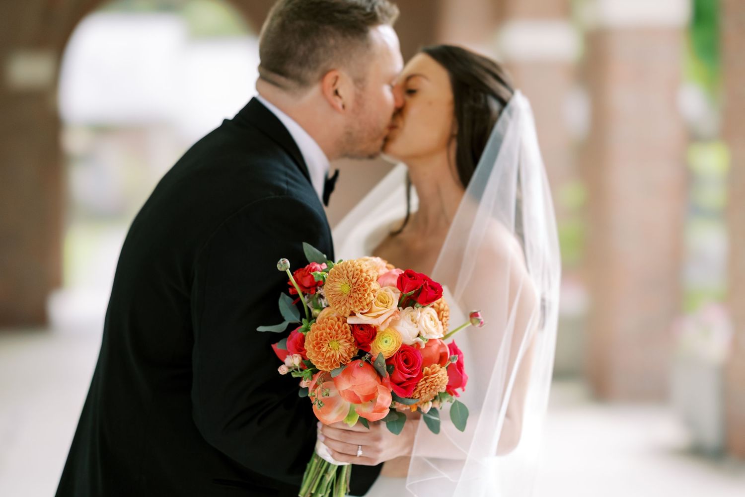 Couple in wedding attire stand under arched brick corridor while bride taps groom's shoulder holding pink bouquet at the Hall of Springs.
