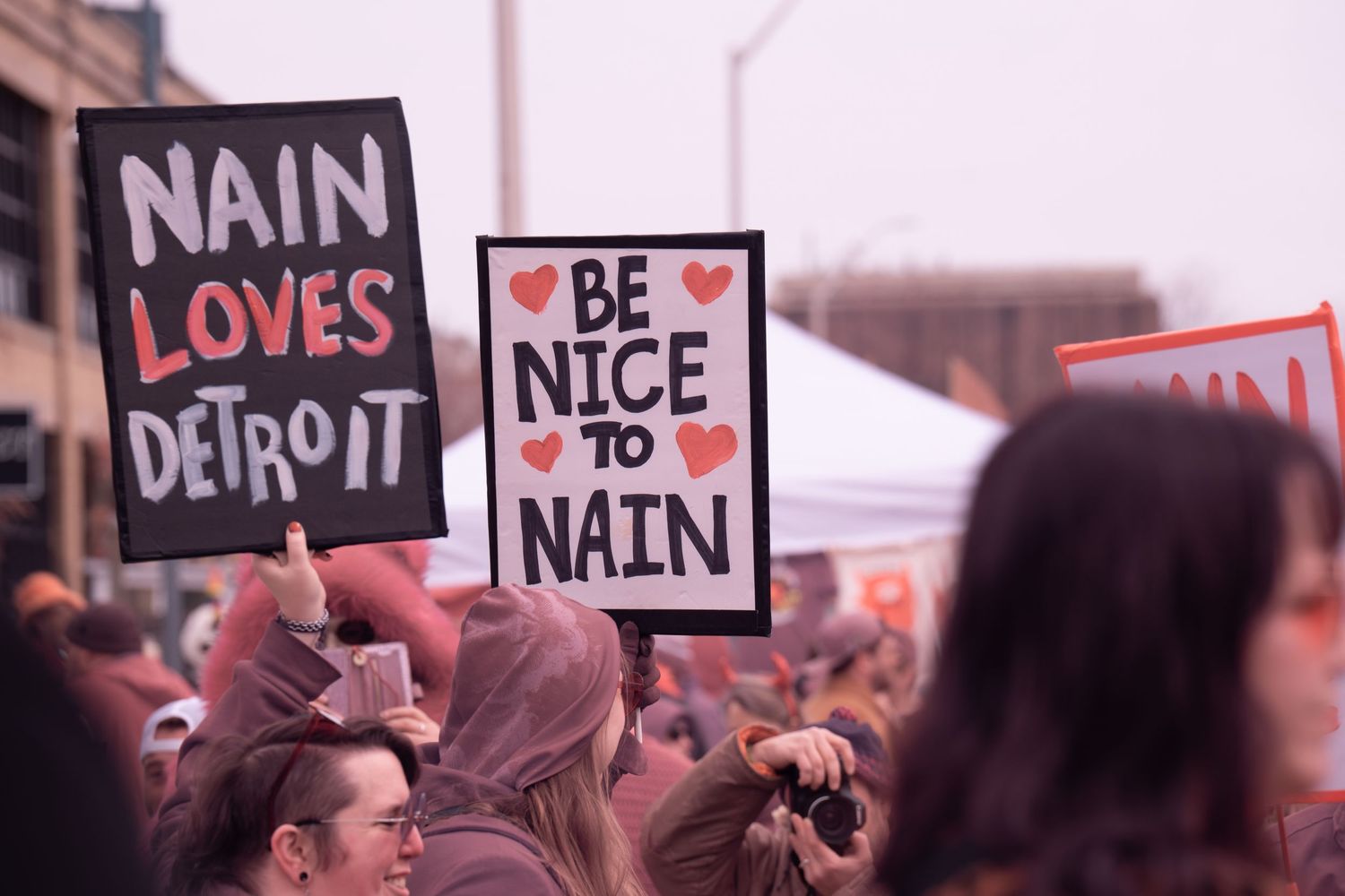 At Nain Rouge event in downtown Detroit, signs reading 'Nain Loves Detroit' and 'Be Nice to Nain' held up in crowd.