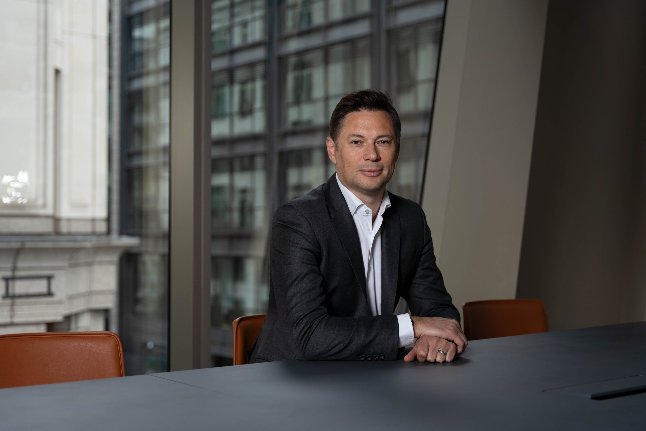 Executive in dark suit sits at conference table near window in modern office setting with professional lighting.