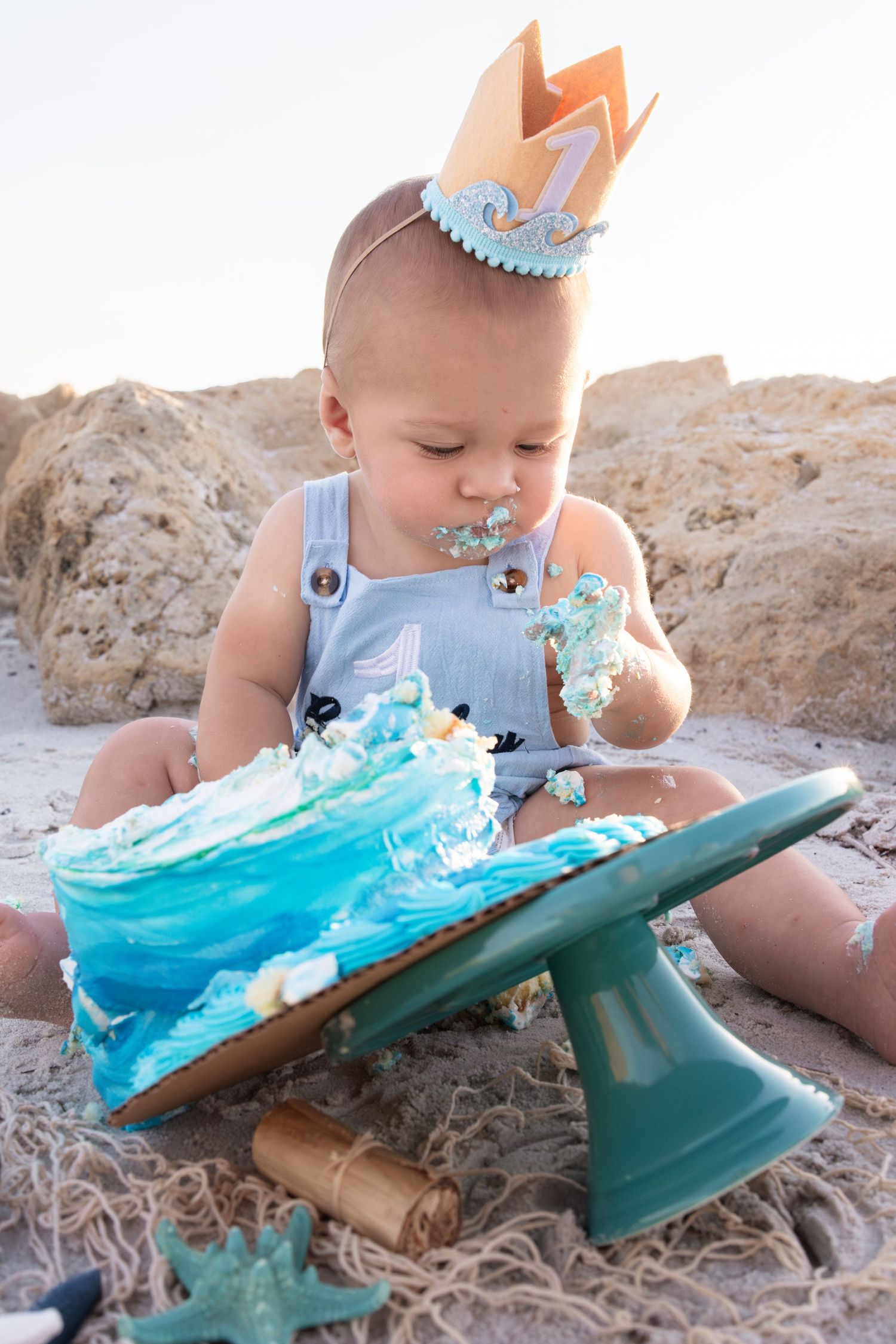 Baby in crown smashes ocean-themed first birthday cake on beach sand.