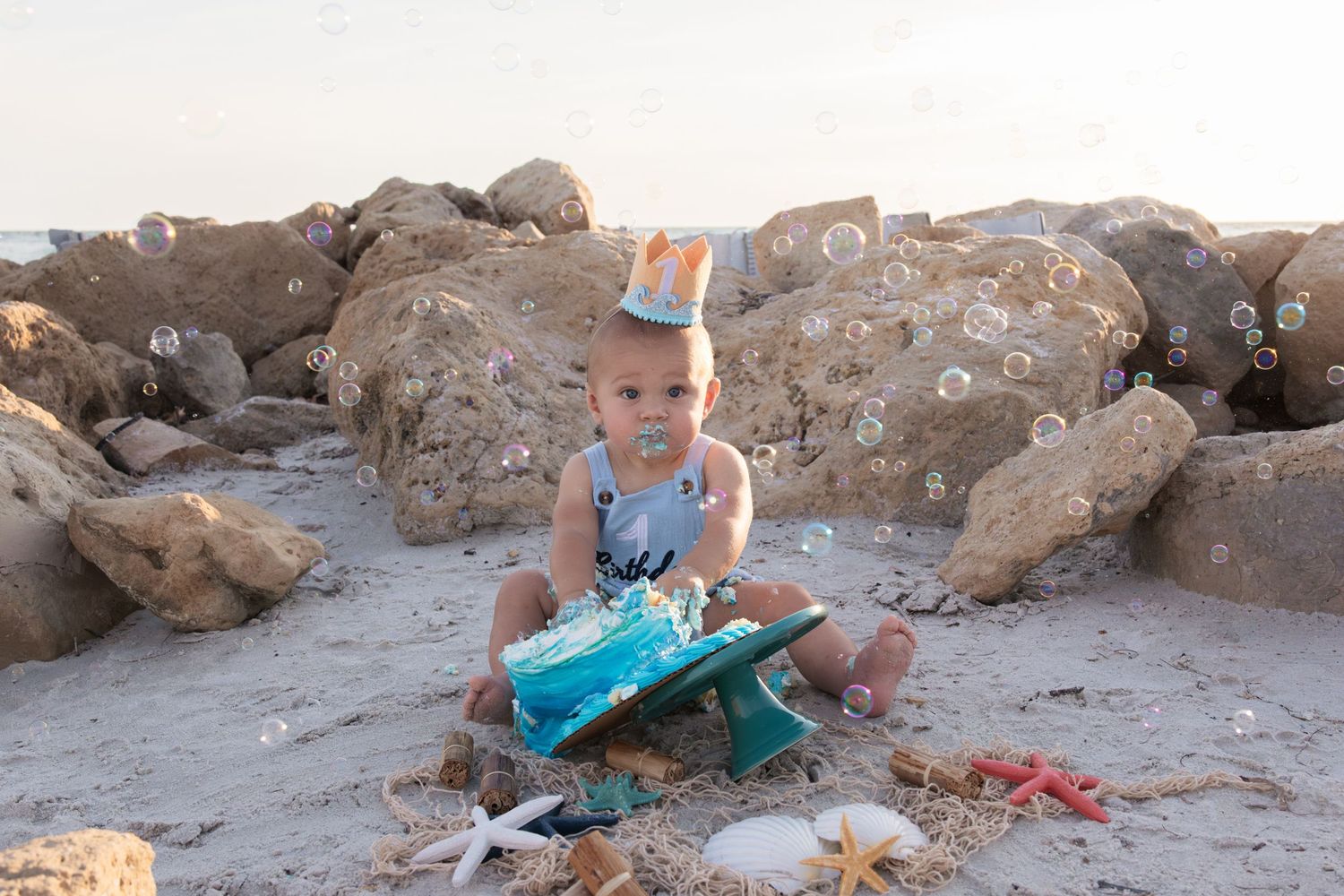 Toddler in birthday crown plays with teal cake on sandy beach surrounded by rocks, shells, and bubbles.