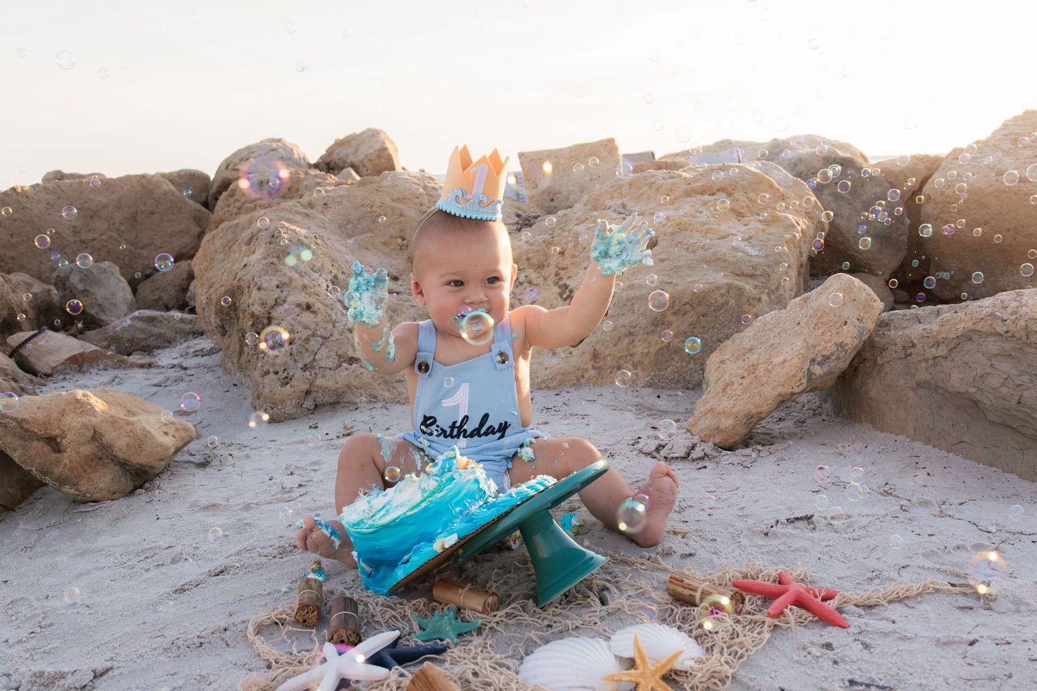 Baby boy in crown smashes first birthday cake on sandy beach surrounded by bubbles and rocks.