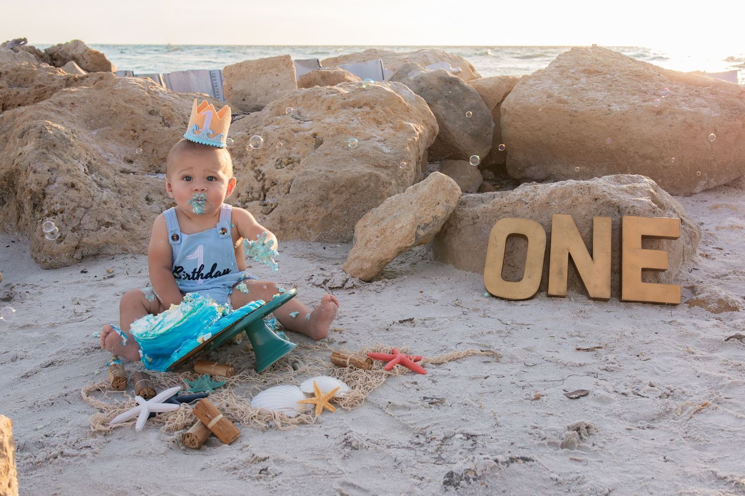 Baby boy in party hat sits on beach sand next to ONE sign, celebrating his first birthday with cake.