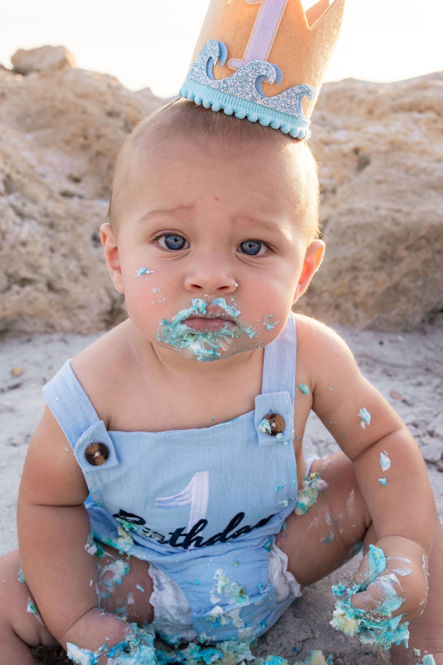 Baby boy in birthday overalls and crown with cake smash frosting on face, celebrating first birthday outdoors.