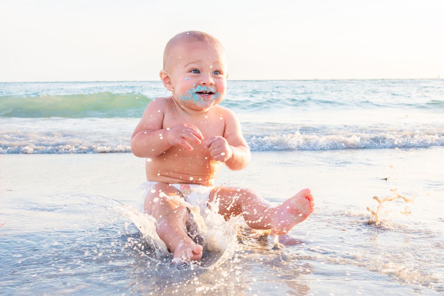 Happy baby splashing in ocean waves on a sunny beach, smiling with a pacifier while sitting in shallow water.