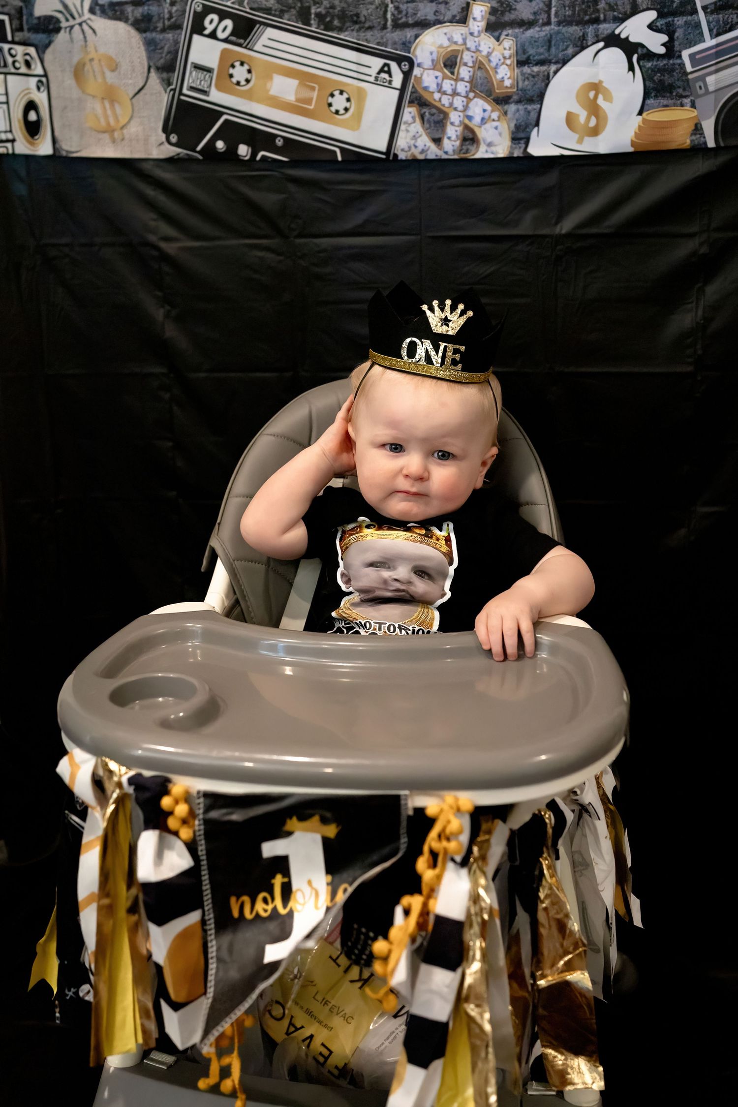 Baby sitting in high chair decorated with gold streamers and wearing a crown for first birthday.