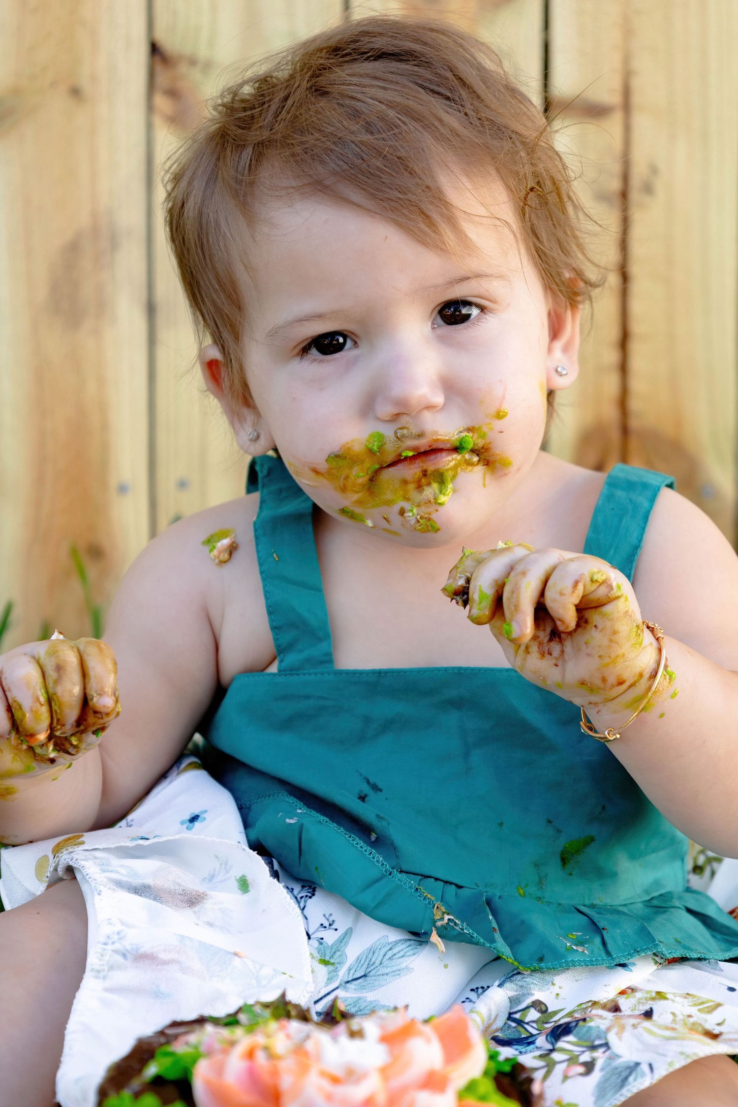 Baby wearing turquoise outfit explores messy cake with hands and face during outdoor photoshoot.