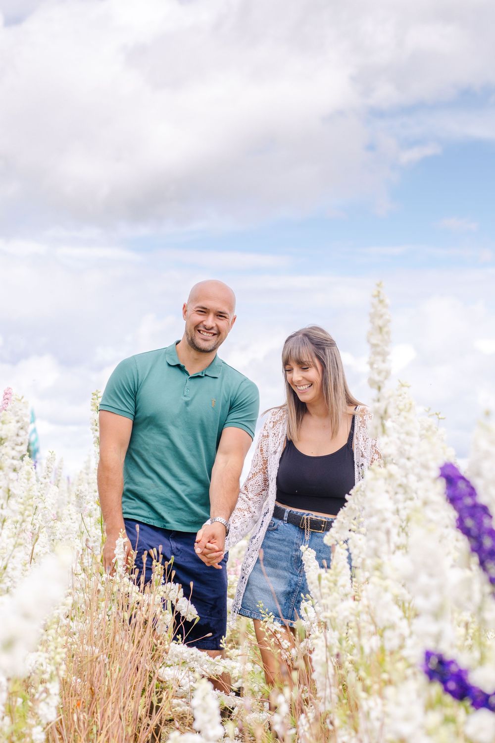 A couple stands together holding hands in a field of white wildflowers on a sunny day with blue skies.